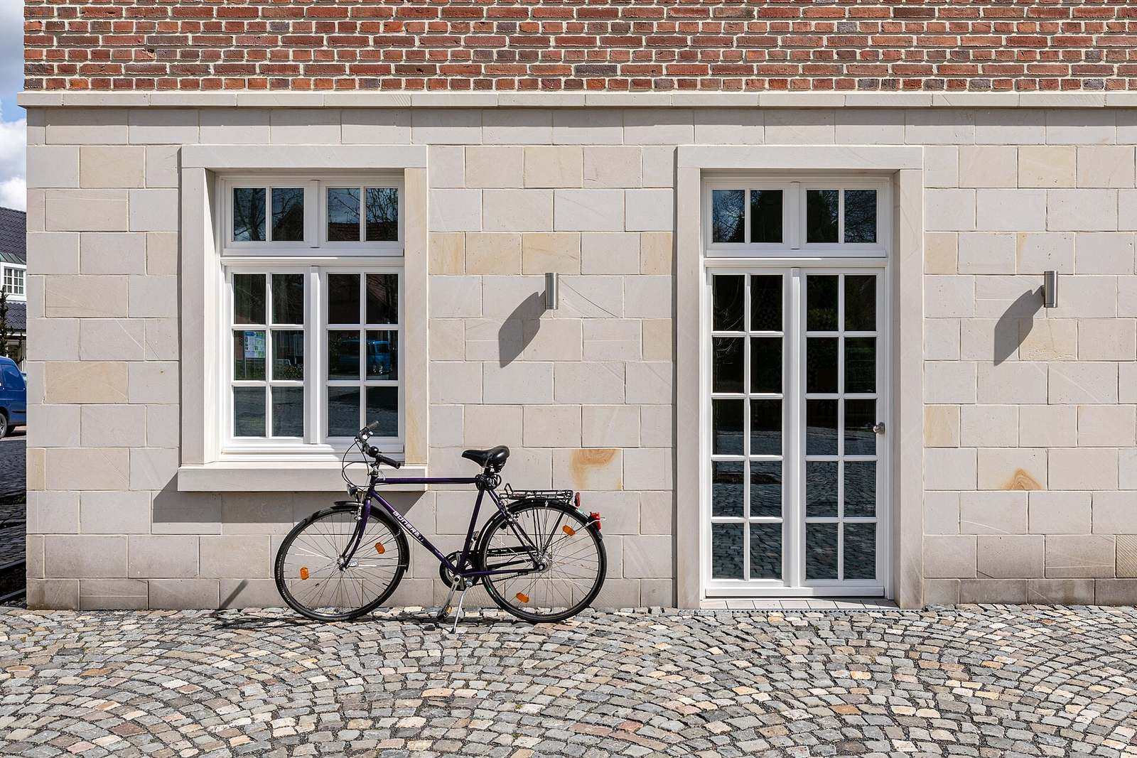 Lichte stenen gevel met een wit raam en een witte glazen dubbele deur, met een fiets op de kasseien