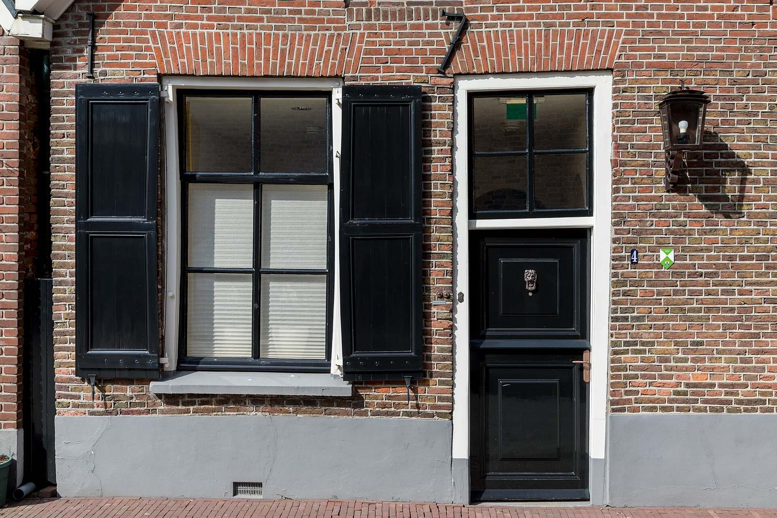 Bakstenen gevel met een donker raam tussen zwarte luiken en een zwarte voordeur met wit kader